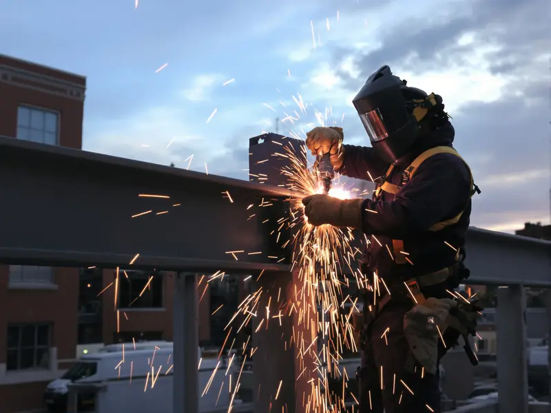 Welder in a safety harness welding a steel I-beam connection at dusk with brick buildings behind